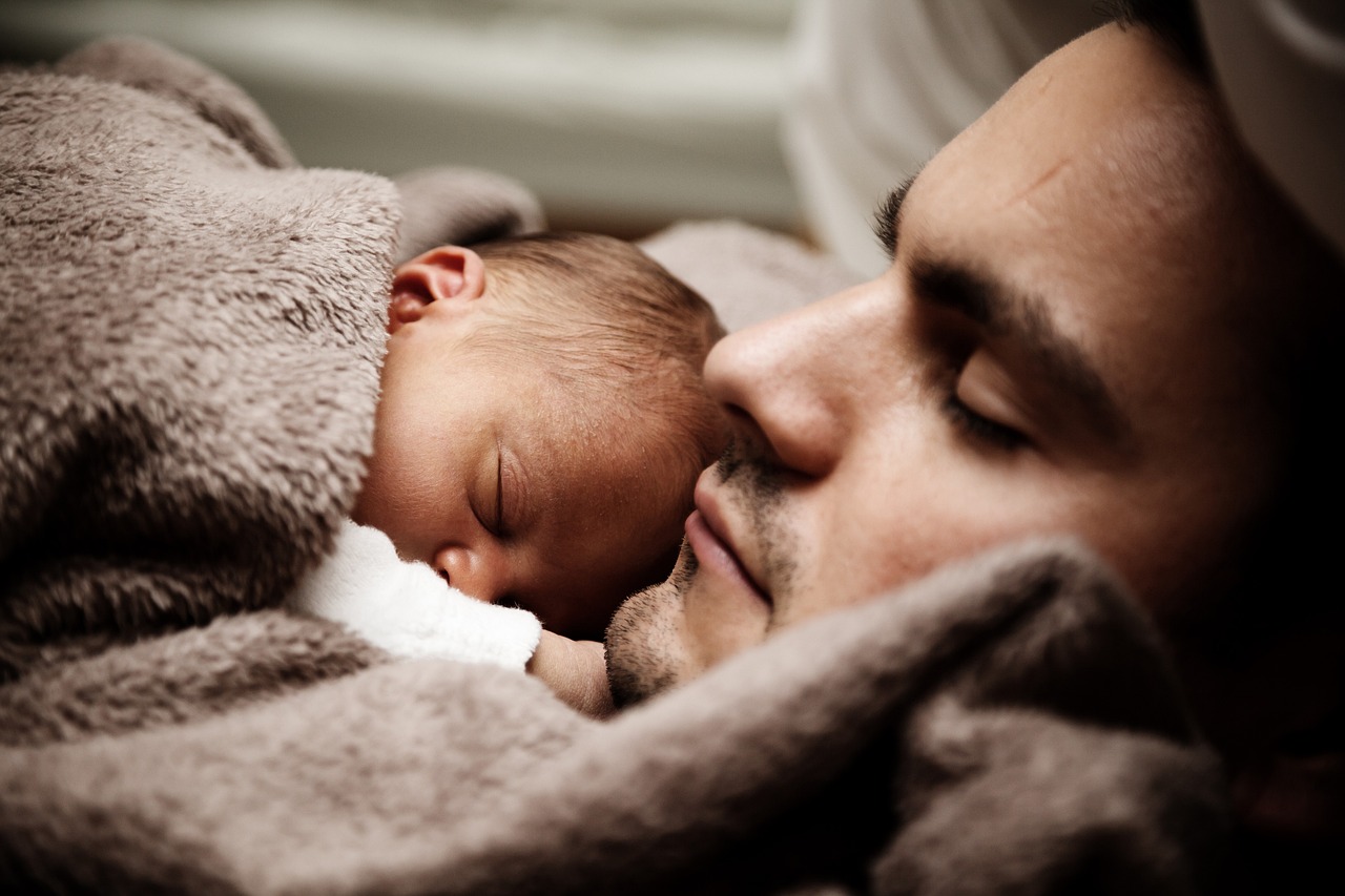 A father and his newborn baby sleeping side by side under a soft taupe blanket, both faces visible in profile.