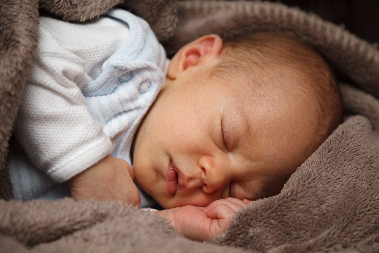 A newborn baby sleeping peacefully on their side, wrapped in a soft taupe blanket and wearing a light blue and white striped outfit.