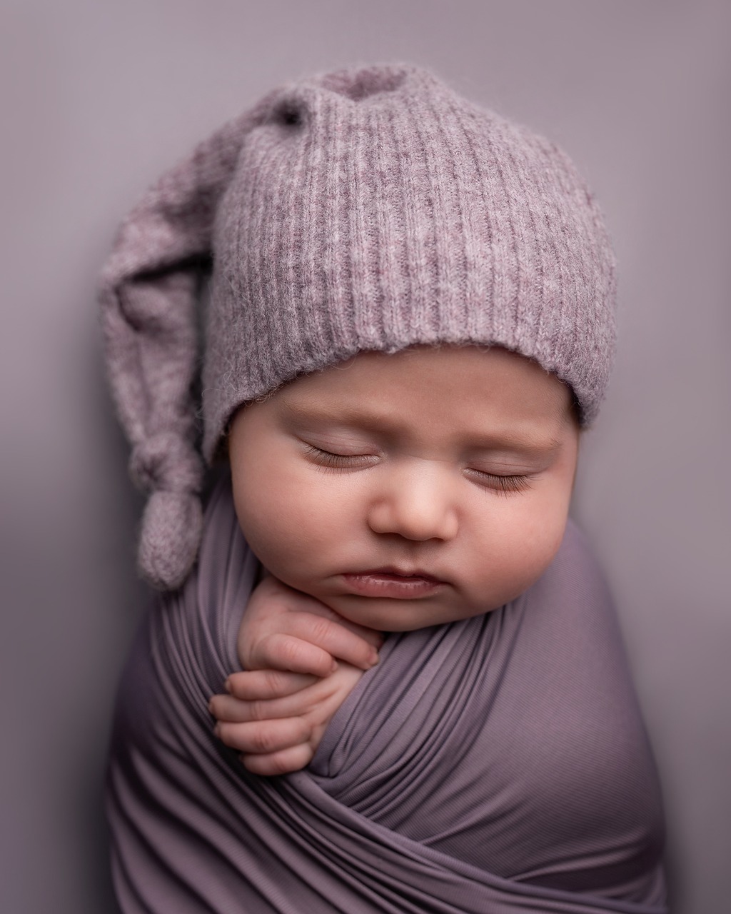 A newborn baby swaddled in soft lilac-grey fabric and wearing a long knit cap, sleeping peacefully.