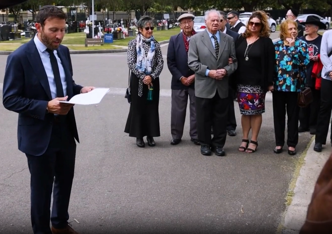 Rabbi Yossi leading a consecration ceremony at a Jewish cemetery, with family gathered.