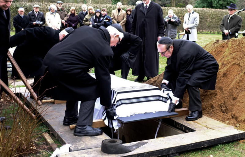 Mourners in dark coats lowering a tallit-draped coffin at a Jewish burial, with family gathered behind.