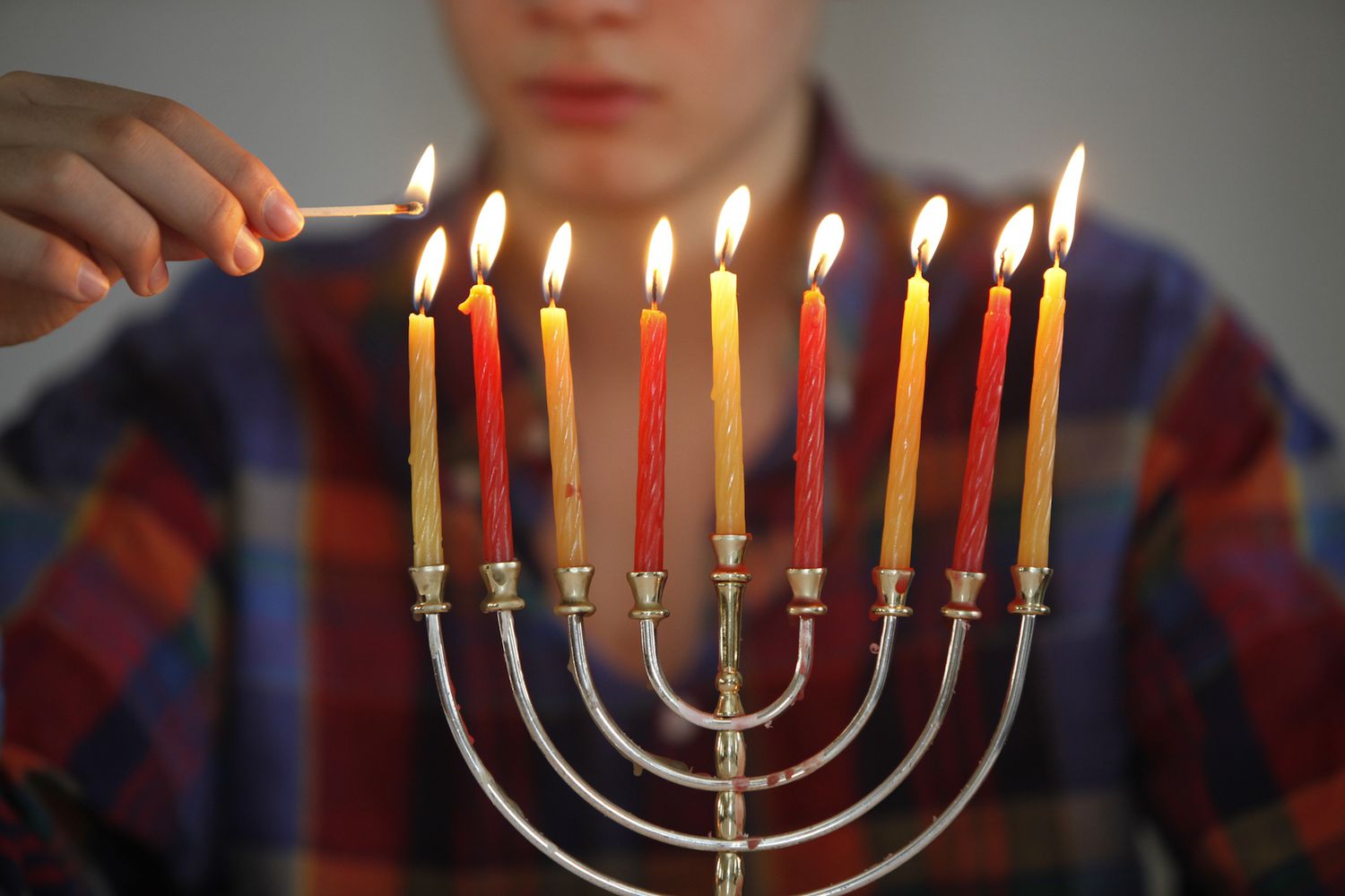 A Hanukkah menorah with its candles lit, a person holding a match behind it